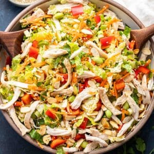 Asian chicken salad in a white salad bowl with wooden serving spoons in the bowl and dressing behind it.