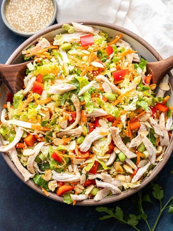 Asian chicken salad in a white salad bowl with wooden serving spoons in the bowl and dressing behind it.