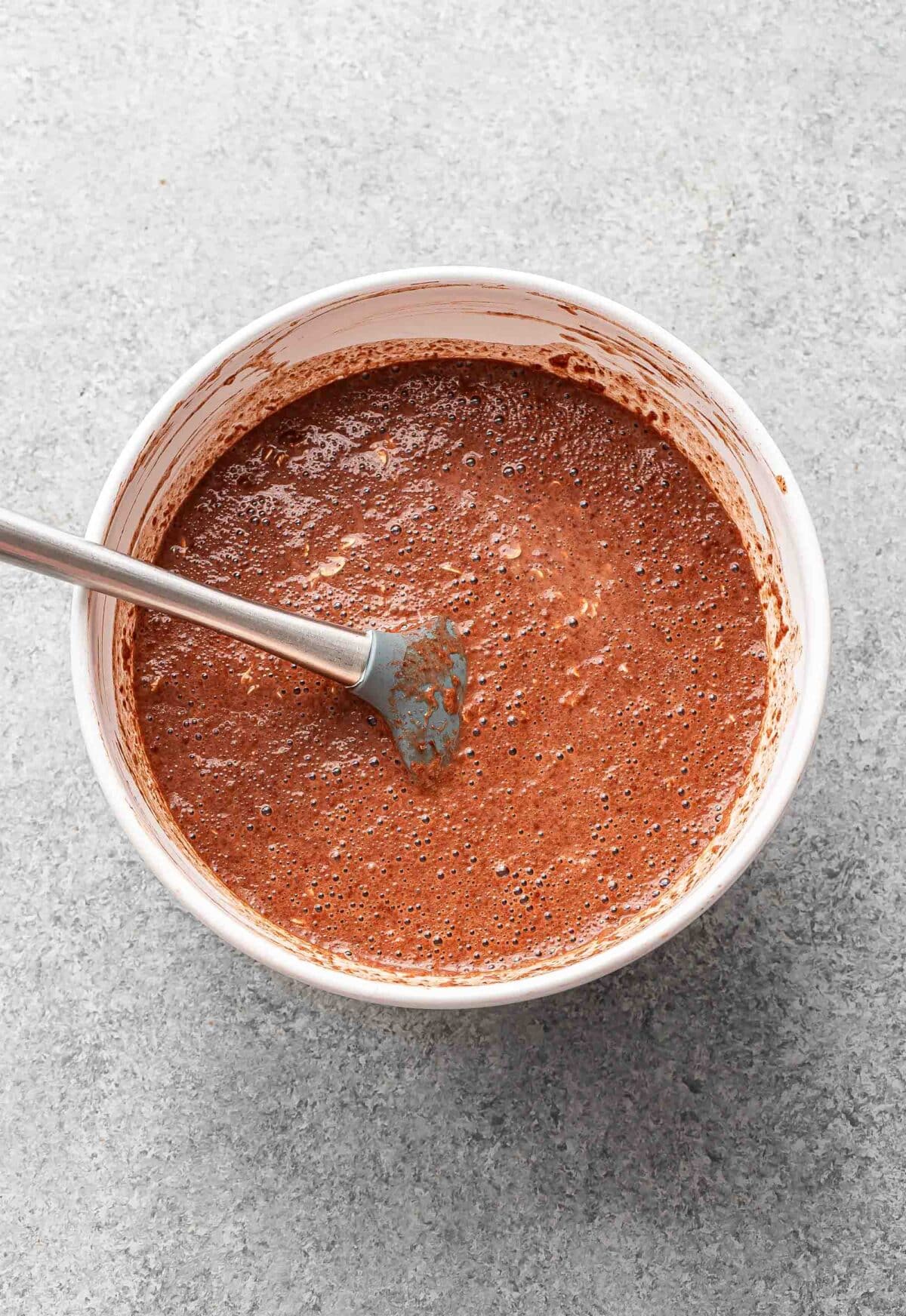 Chocolate baked oatmeal mixture in a white mixing bowl with a rubber spatula before it's been baked.