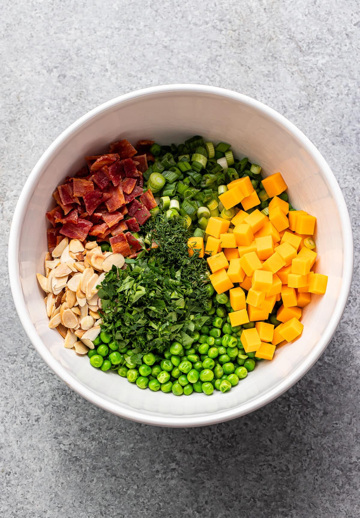 Peas, cheddar cheese, herbs, green onions, bacon, and almonds in a white bowl.