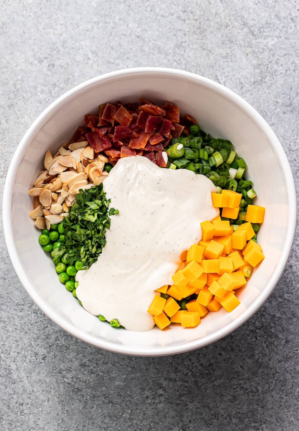 Peas, cheddar cheese, herbs, green onions, bacon, and almonds in a white bowl with creamy dressing on top before it's been mixed together.