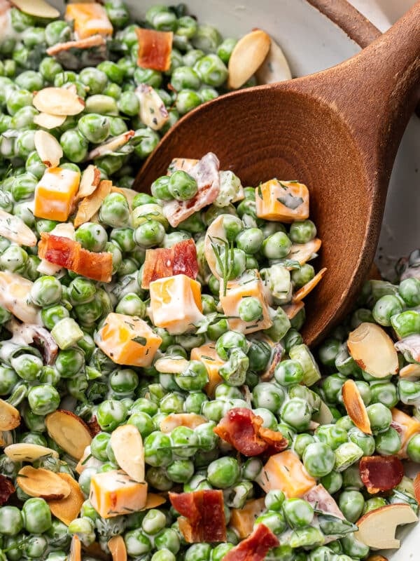 A wooden serving spoon scooping up pea salad with cheddar cheese and bacon out of a serving bowl.