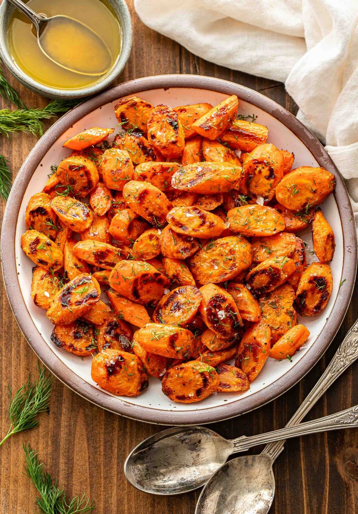 Roasted carrots with honey butter and dill on a white plate. A bowl of honey butter is behind the plate and two serving spoons in front of the plate.