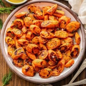 Roasted carrots with honey butter and dill on a white plate. A bowl of honey butter is behind the plate and two serving spoons in front of the plate.