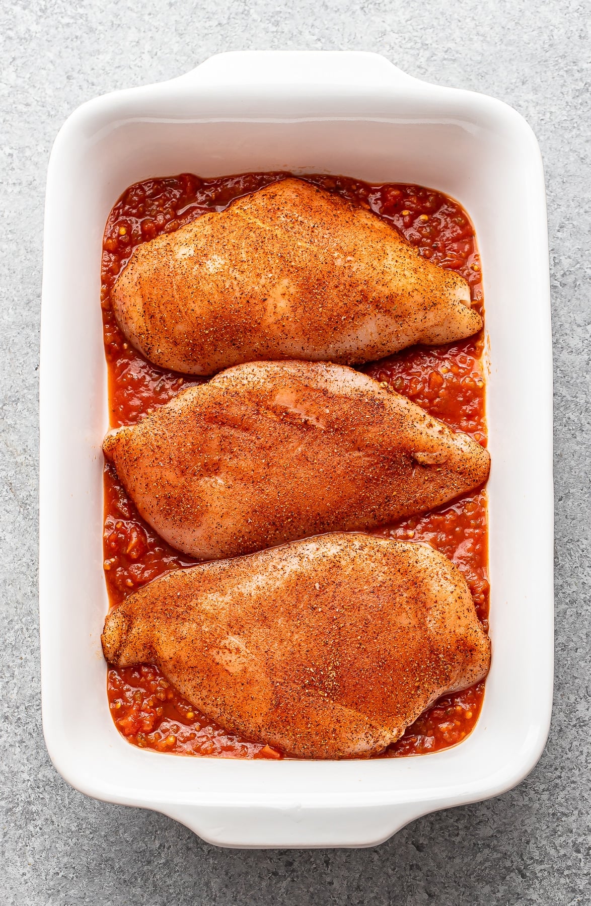 Three seasoned chicken breasts in a casserole dish on top of salsa.