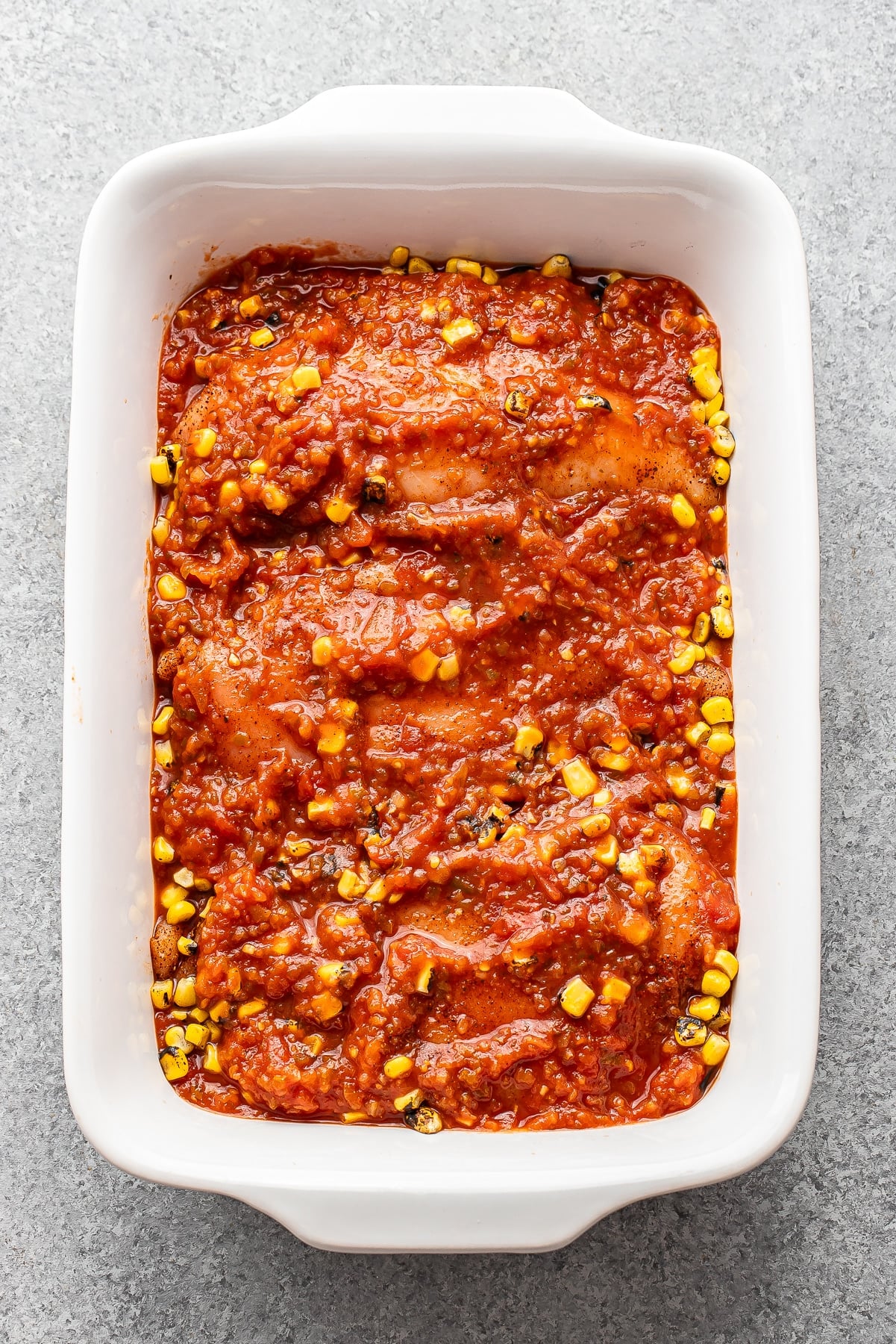 A casserole dish with three chicken breasts, corn, and salsa before it's been baked.