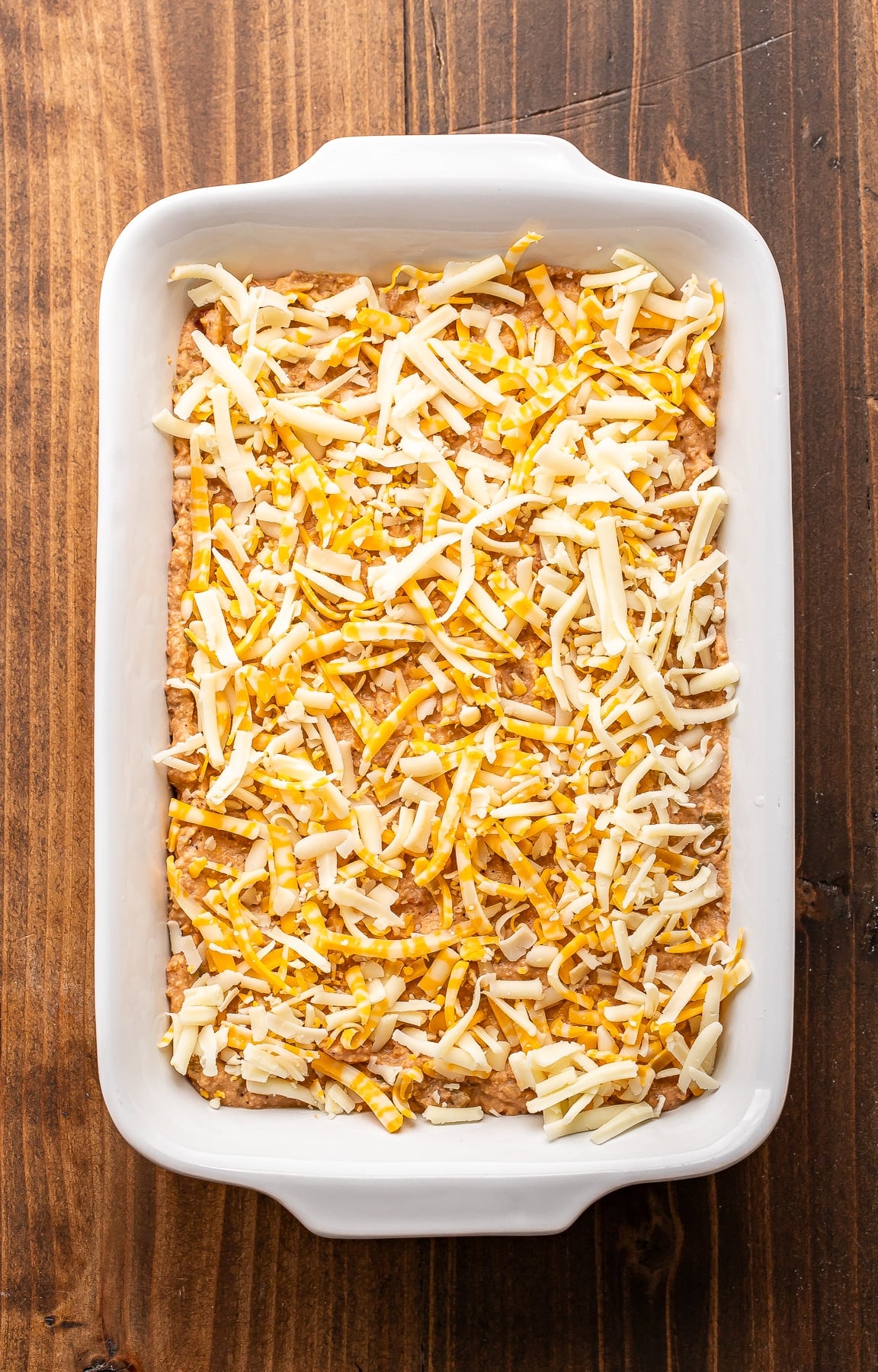 Bean dip before it's been baked topped with shredded cheese in a white baking dish.