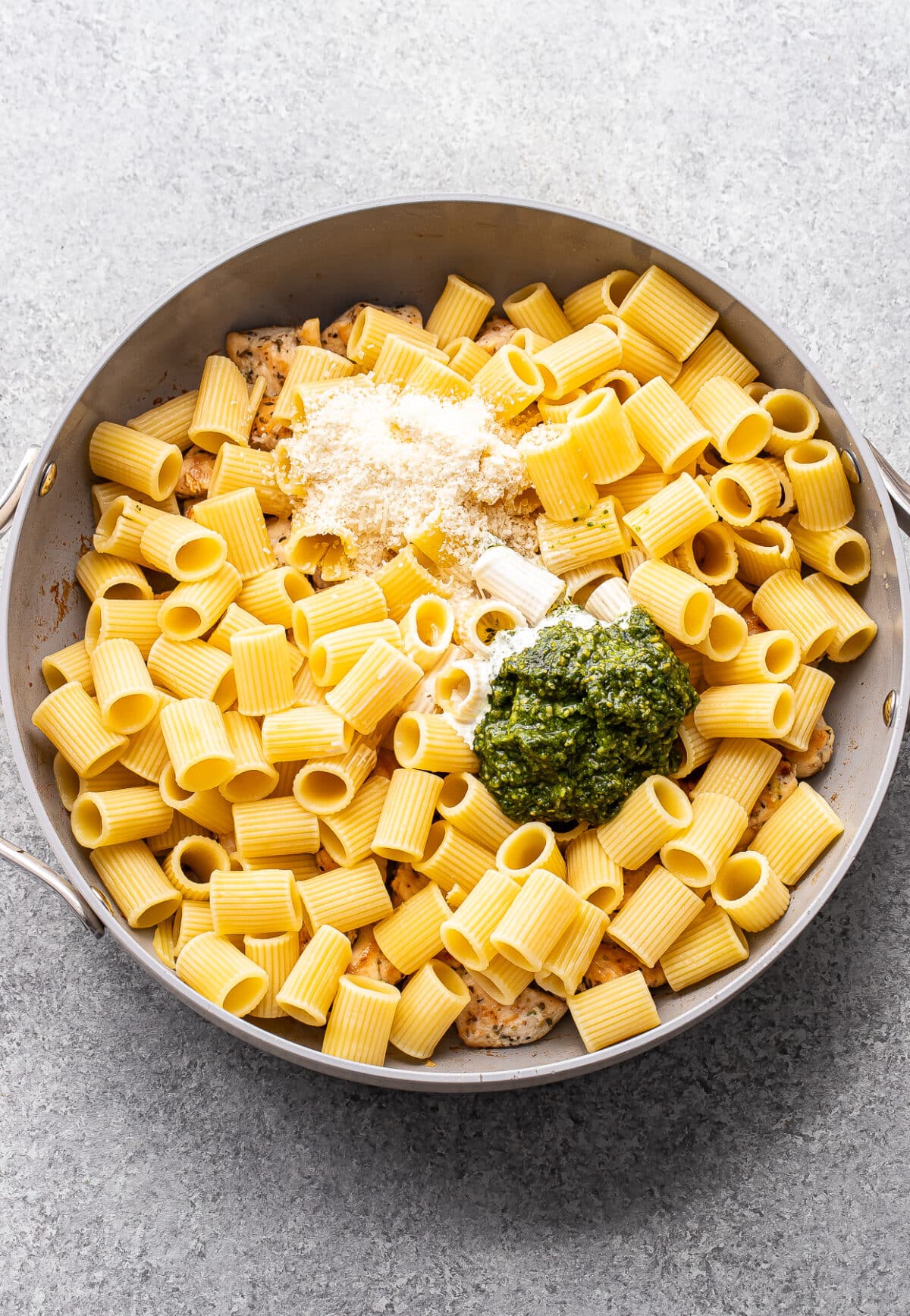 Pasta, chicken, pesto, cream, and parmesan cheese in a pan before being mixed together.