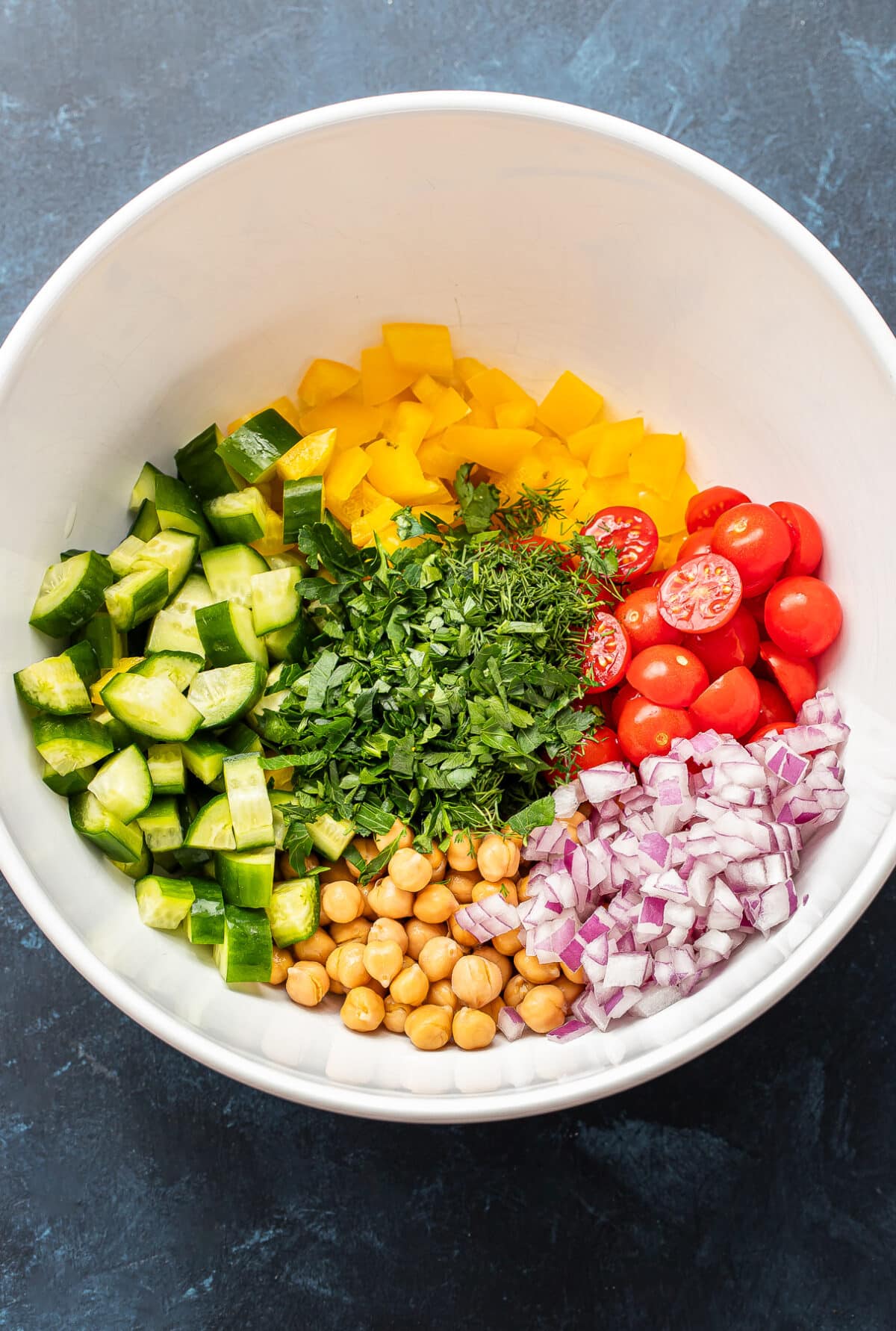 Peppers, tomatoes, red onion, cucumber, chickpeas, and herbs in a white mixing bowl.