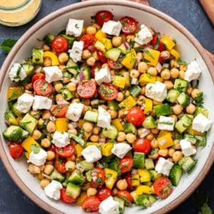 Chickpea salad with tomatoes, feta, cucumbers, peppers, and herbs in a white salad bowl with a jar of lemon vinaigrette behind it.