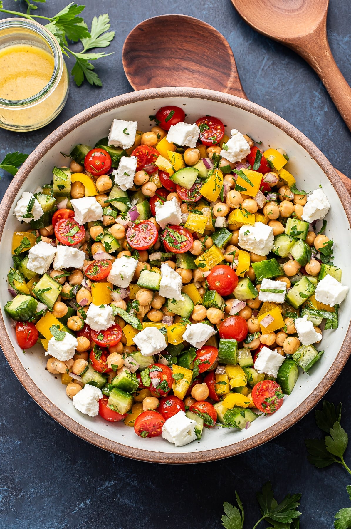 Chickpea salad with tomatoes, feta, cucumbers, peppers, and herbs in a white salad bowl with a jar of lemon vinaigrette behind it.