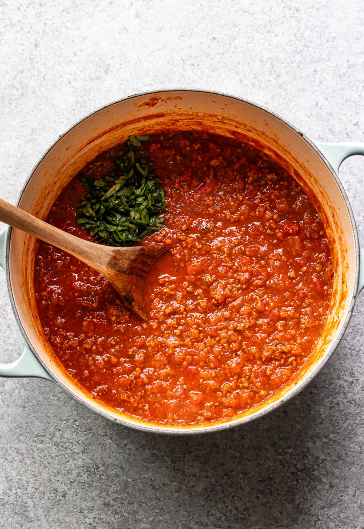 Ground beef spaghetti sauce in a pot with a wooden spoon and chopped basil on top.
