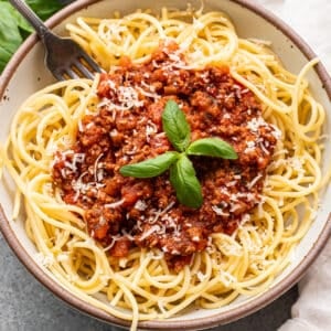 Spaghetti meats sauce garnished with basil leaves on top of spaghetti noodles in a white bowl with a fork.