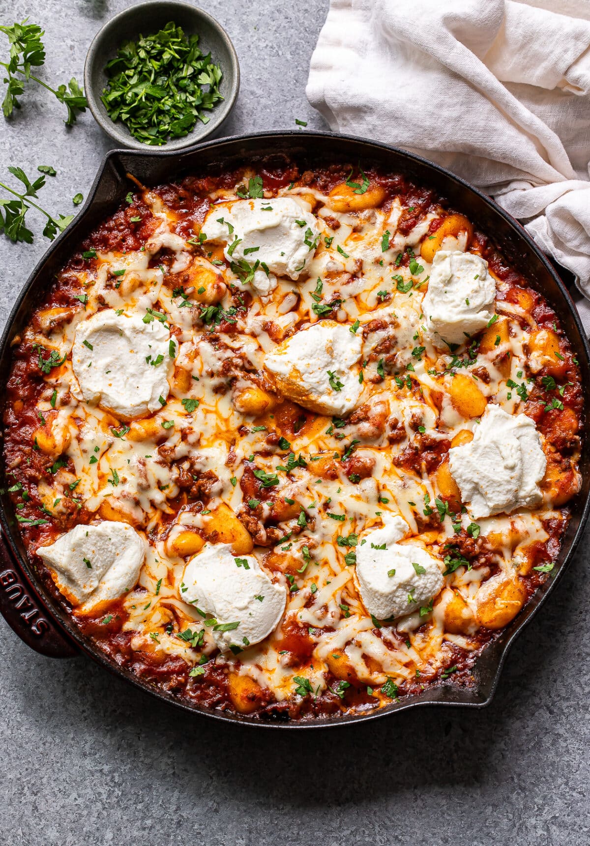 Baked Italian ground beef and gnocchi in a skillet. A bowl of chopped parsley is behind the skillet.