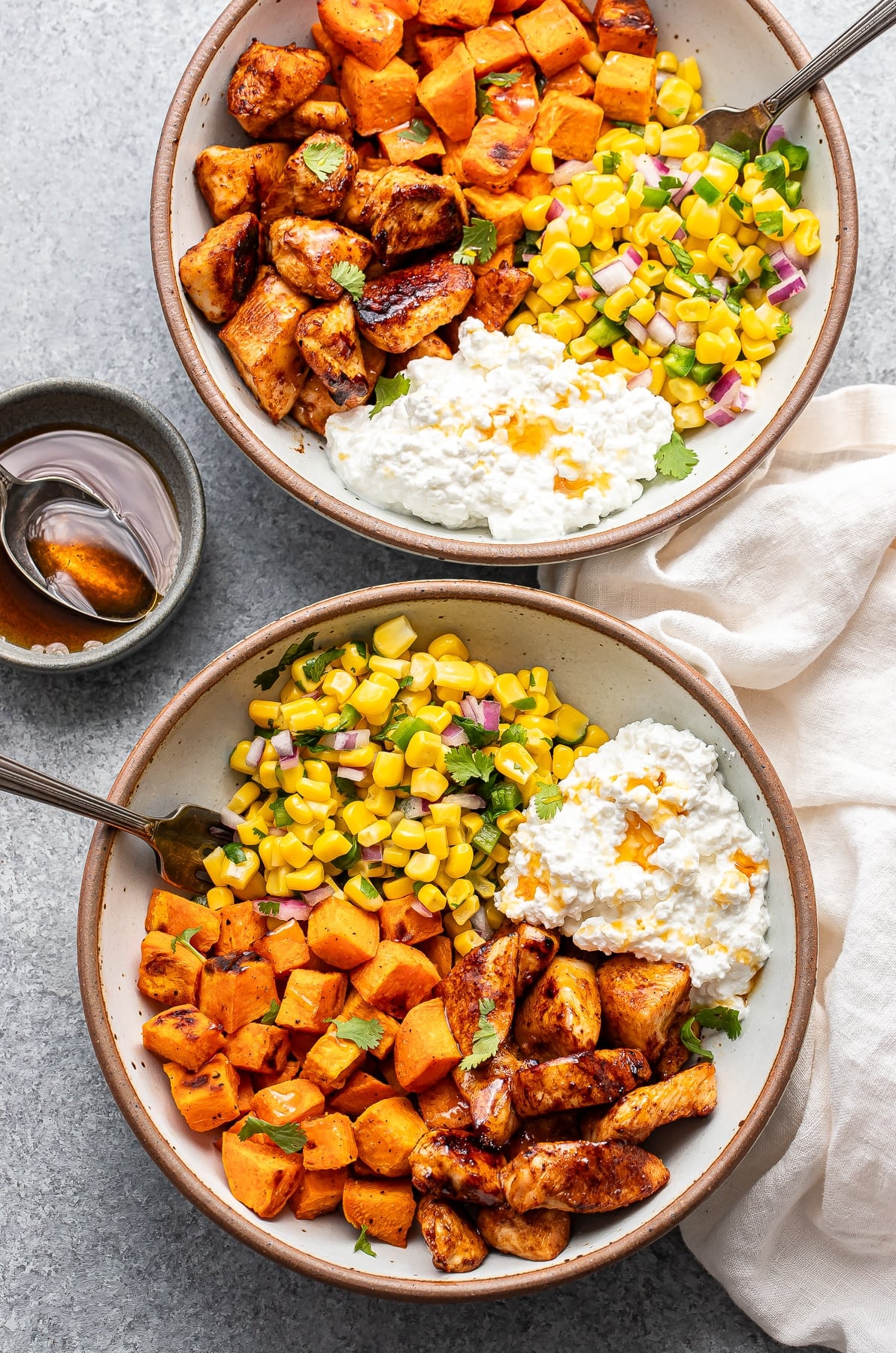 Two hot honey chicken bowls with cottage cheese, sweet potatoes, and corn salsa in white bowls with forks.