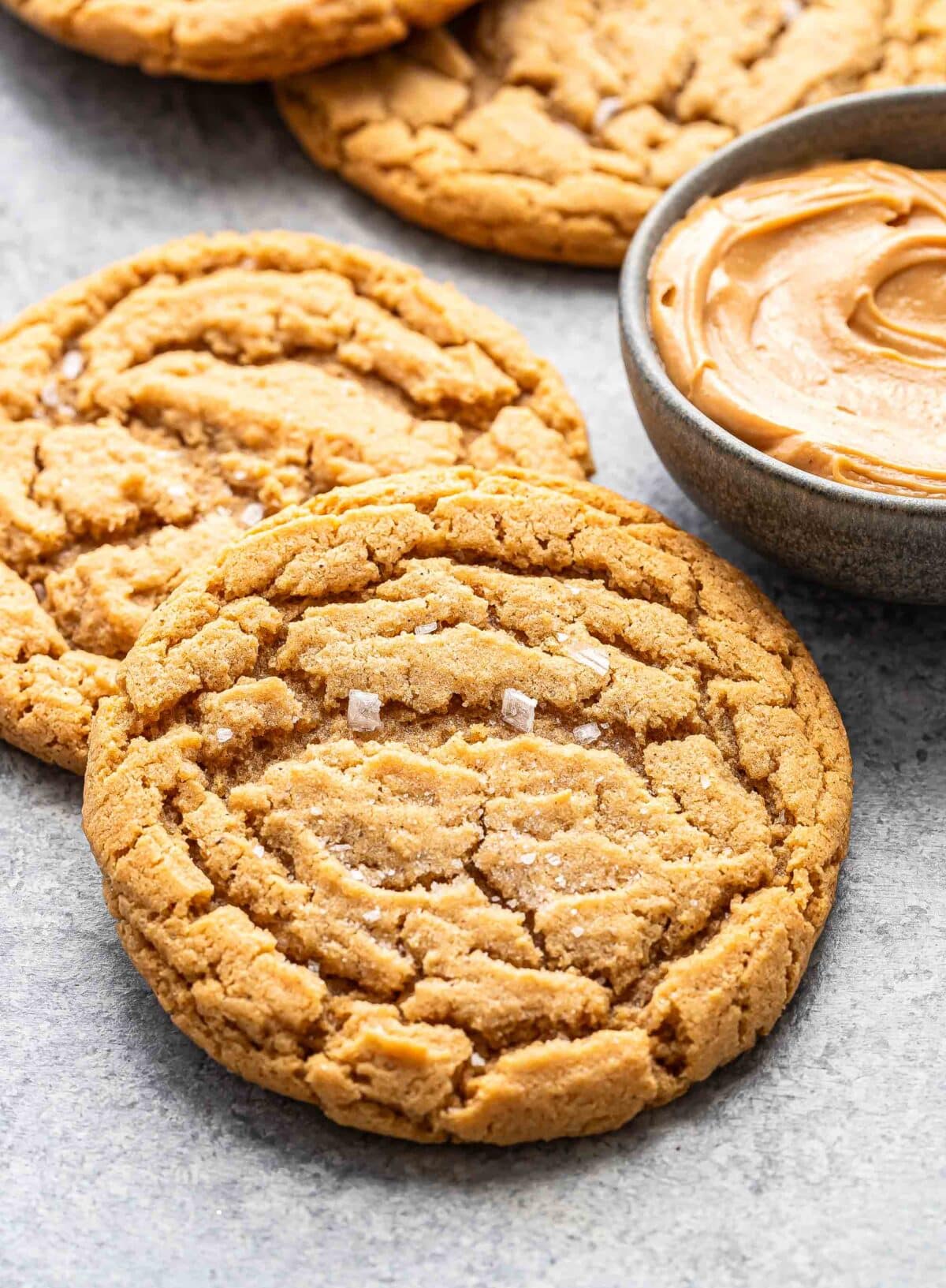 Two peanut butter cookies overlapping each other next to a bowl of peanut butter.