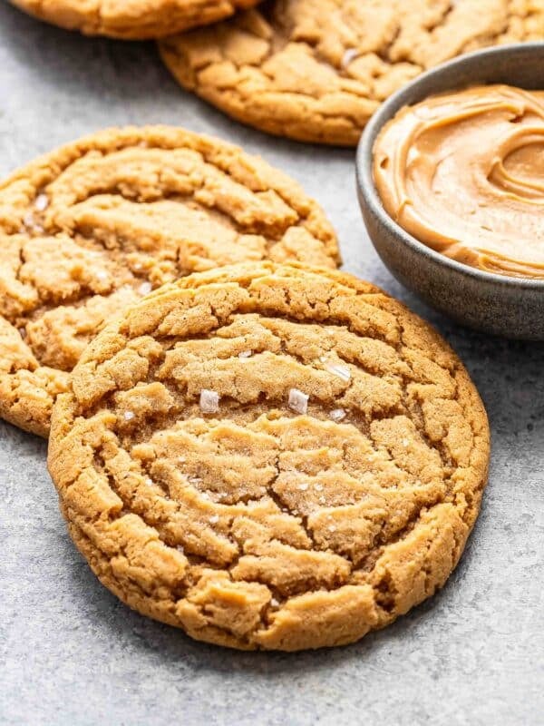 Two peanut butter cookies overlapping each other next to a bowl of peanut butter.