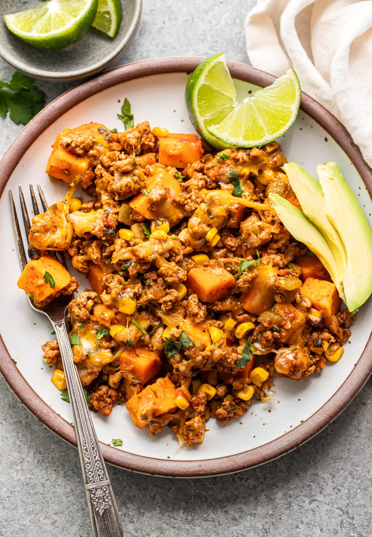 Southwest ground turkey and sweet potatoes on a white plate with a fork, avocado slices, and lime wedges.