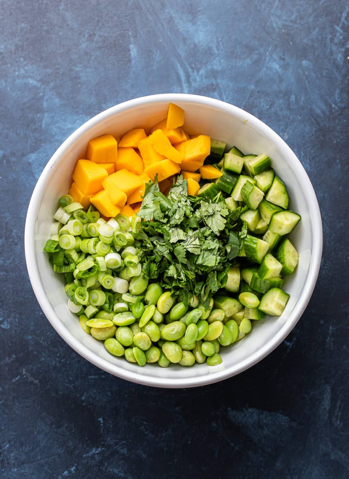 Diced mango, cucumber, green onions, edamame and cilantro in a white mixing bowl.