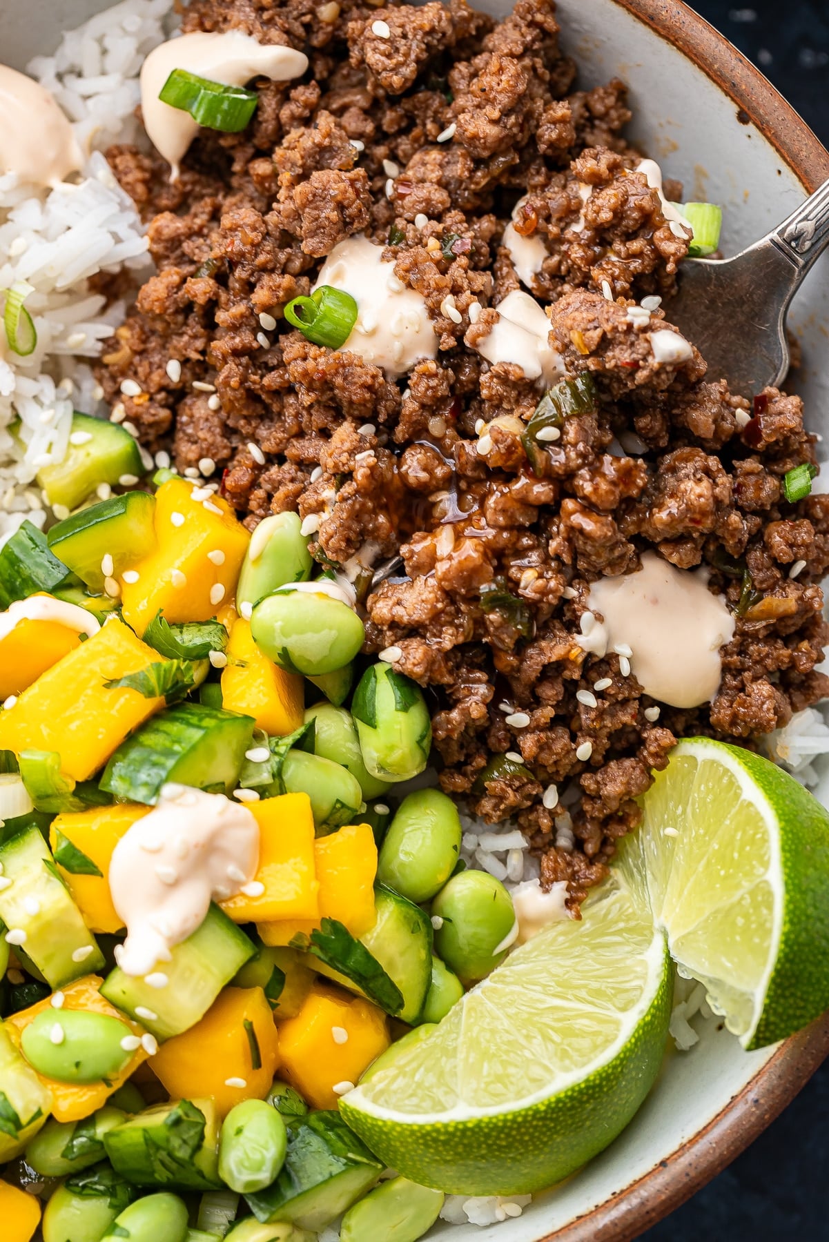 Sweet chili ground beef, mango cucumber salad, rice, and lime wedges in a white bowl with a fork.