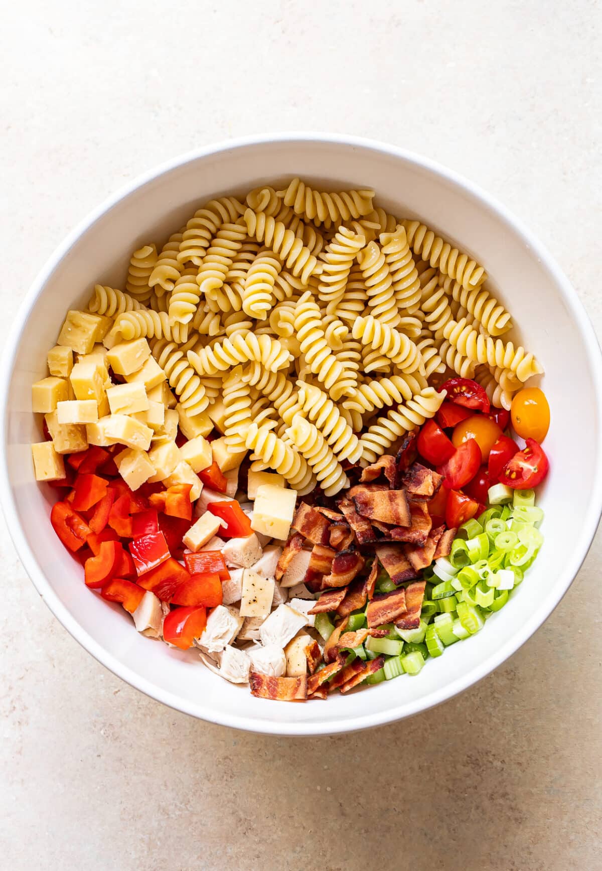 A white bowl filled with uncooked pasta spirals, diced cheese, halved cherry tomatoes, chopped red bell pepper, cooked chicken pieces, crispy bacon, and sliced green onions, arranged in separate sections.