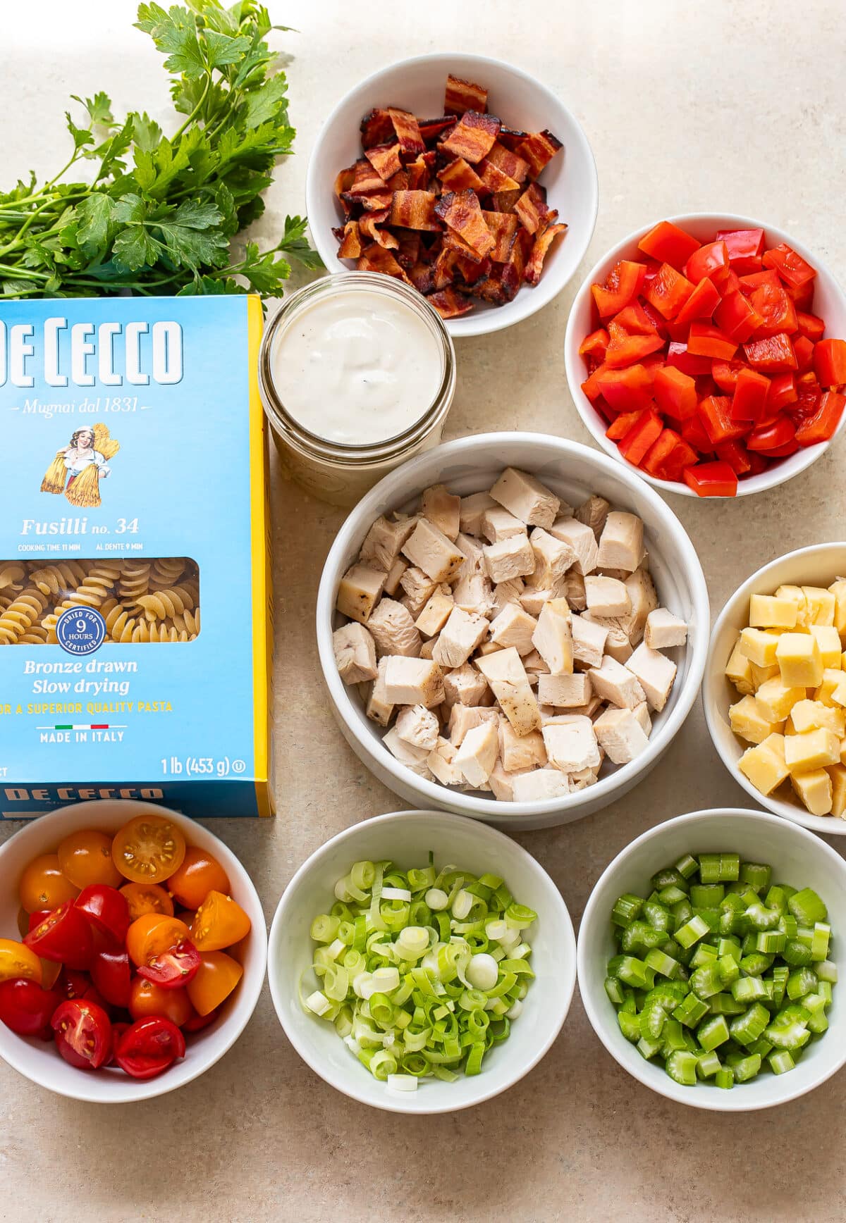 Bowls of diced cooked chicken, bacon, red bell pepper, cheddar cheese, celery, green onions, and cherry tomatoes are arranged around a box of fusilli pasta, a jar of creamy dressing, and fresh parsley on a countertop.