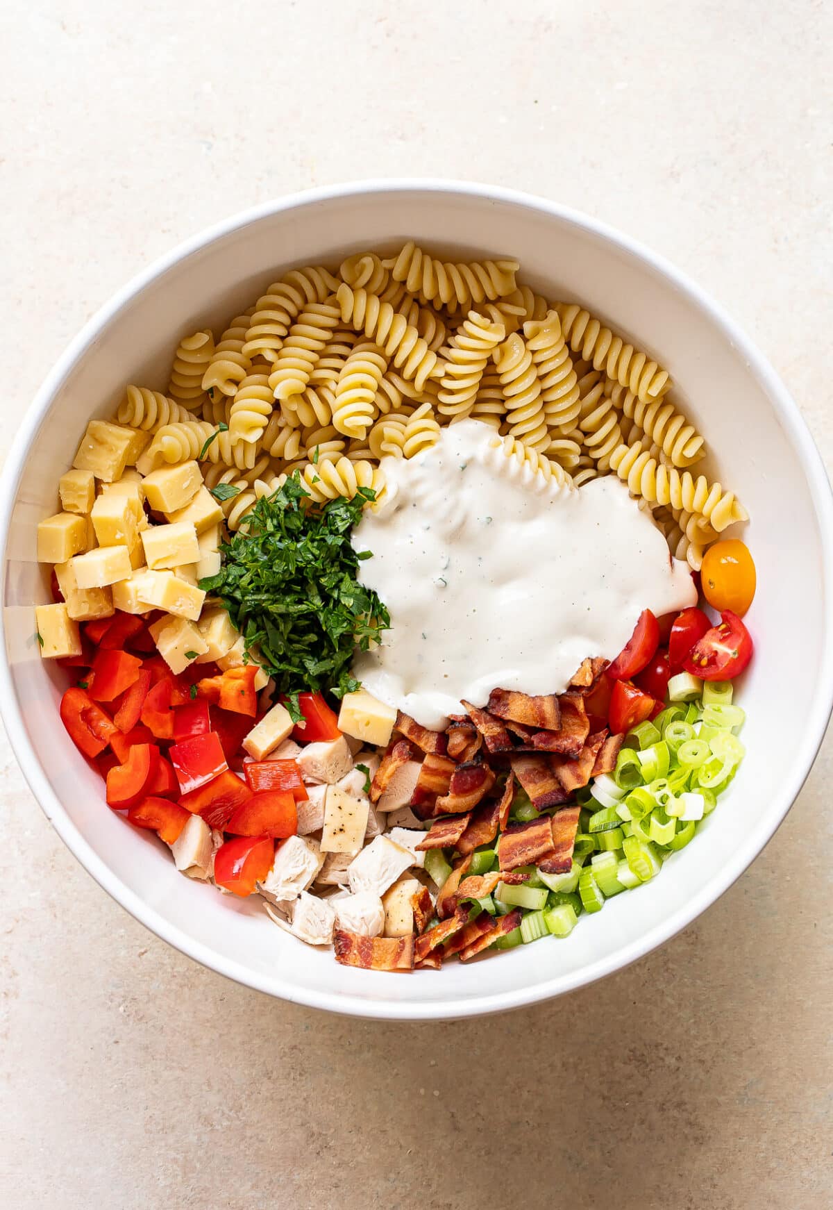 A white bowl filled with rotini pasta, diced cheddar cheese, chopped parsley, ranch dressing, sliced cherry tomatoes, celery, green onions, red bell pepper, cooked bacon, and grilled chicken pieces.
