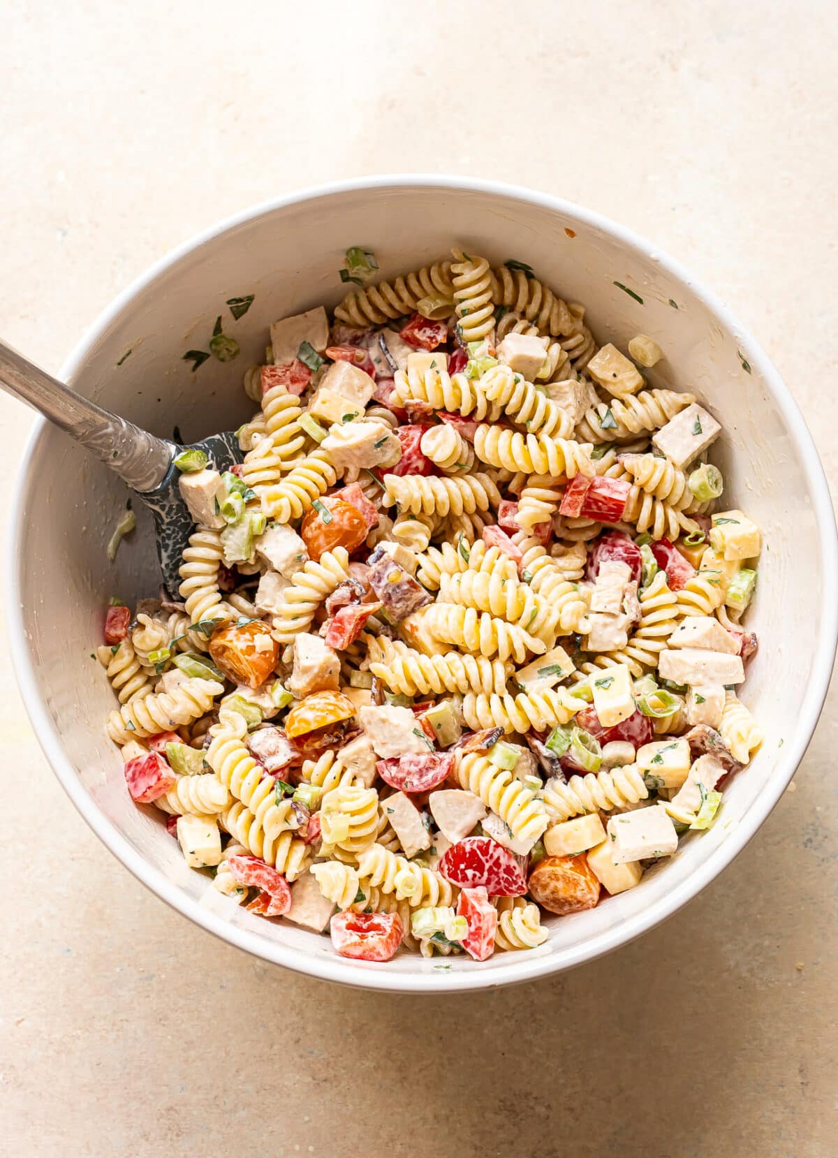 A white bowl filled with rotini pasta salad, featuring diced chicken, cherry tomatoes, green onions, and cubes of cheese, all mixed with a creamy dressing. A serving spoon rests in the bowl.