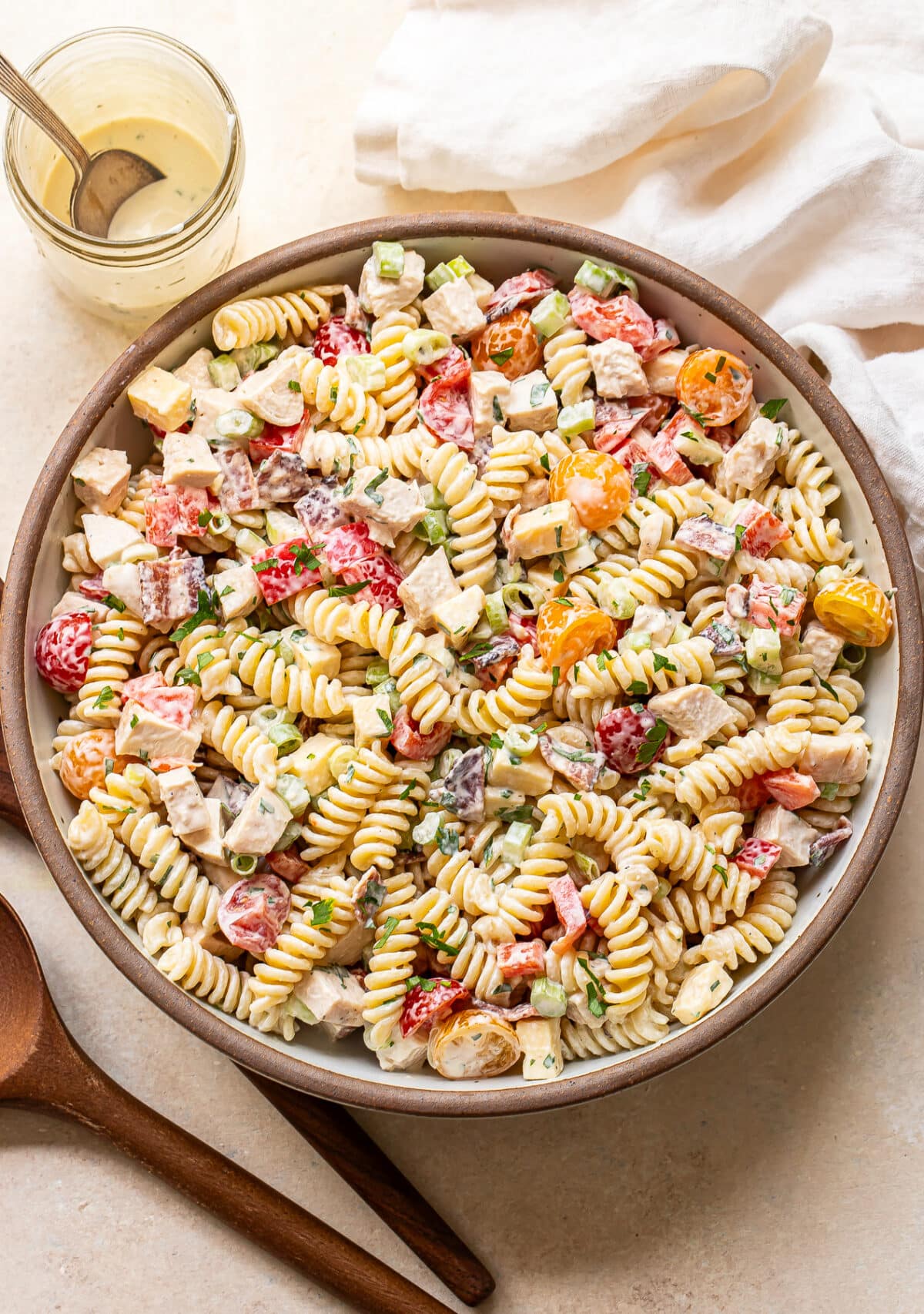 A large bowl of rotini pasta salad with diced chicken, cherry tomatoes, red onion, cucumber, and herbs, all mixed with a creamy dressing. A jar of dressing, a white napkin, and wooden utensils are beside the bowl.