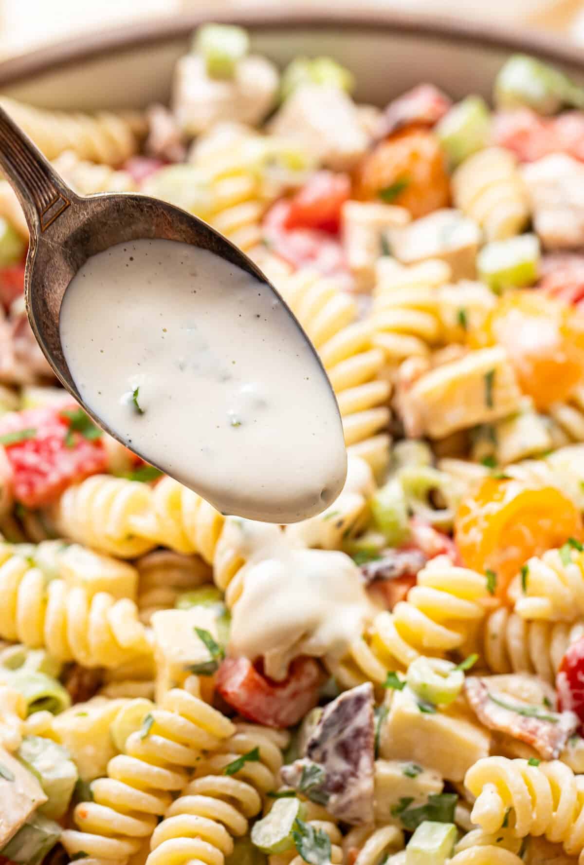 A close-up of a spoon pouring creamy dressing over a bowl of rotini pasta salad with chopped vegetables and herbs.