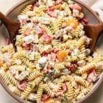 A bowl of creamy pasta salad with rotini, chopped vegetables, and herbs is being served with wooden utensils. A glass of dressing and a white napkin are nearby.