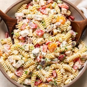 A bowl of creamy pasta salad with rotini, chopped vegetables, and herbs is being served with wooden utensils. A glass of dressing and a white napkin are nearby.