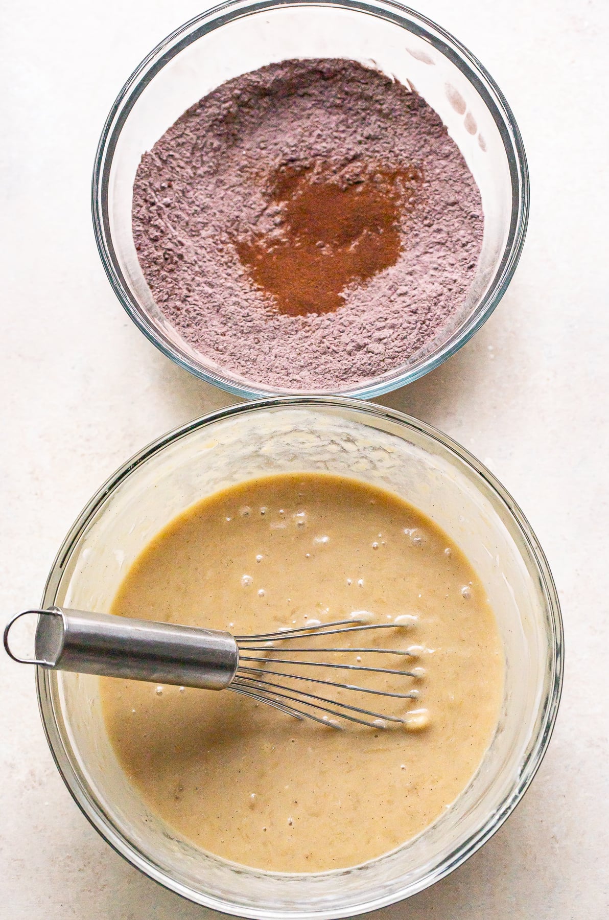 Wet ingredients for chocolate banana bread in a glass mixing bowl with a whisk. Dry ingredients in a separate glass bowl.