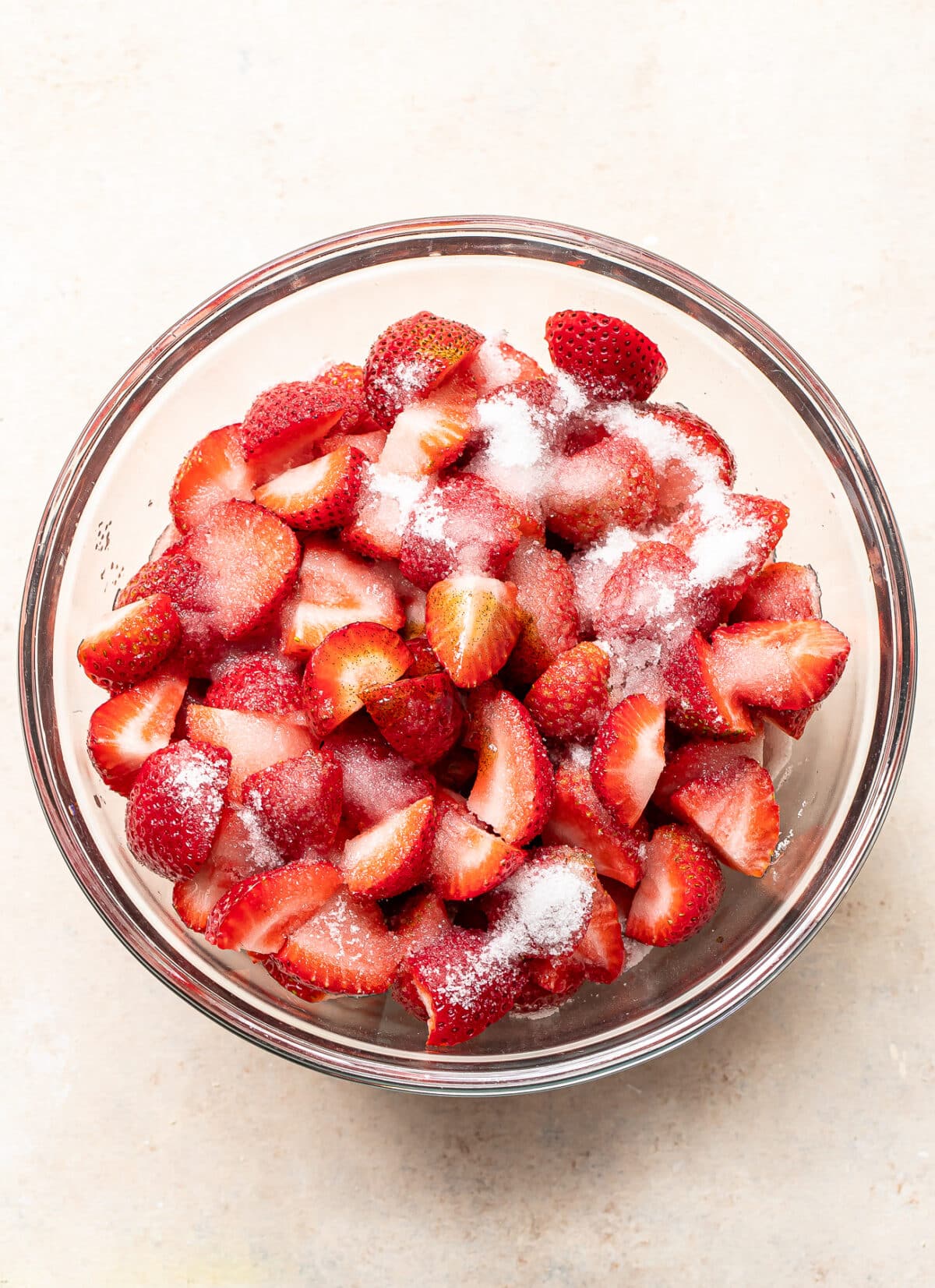 A glass bowl filled with sliced strawberries sprinkled with granulated sugar, placed on a light-colored surface.