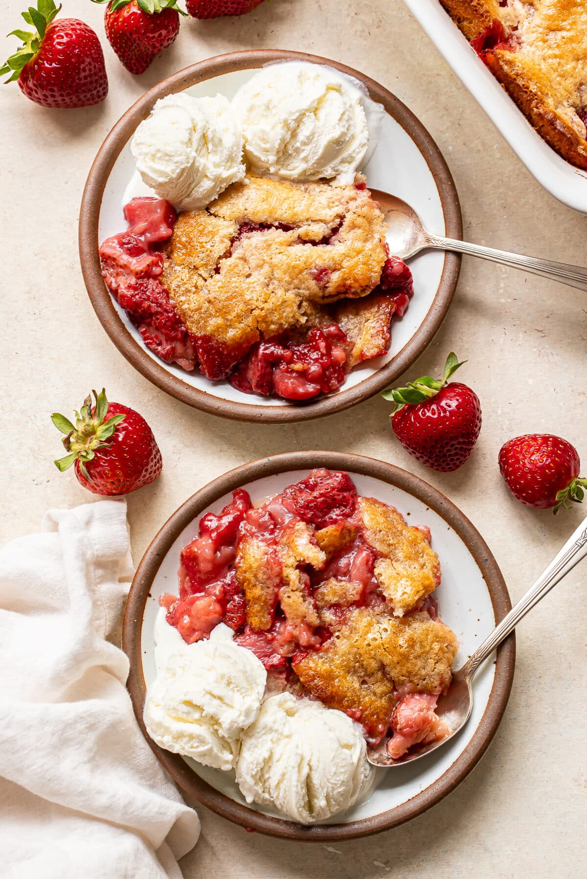 Two plates with servings of strawberry cobbler and scoops of vanilla ice cream. Fresh strawberries are scattered nearby on the light-colored surface, with a white napkin to the side.