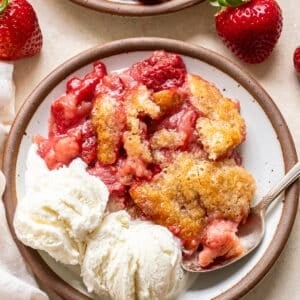 A plate with strawberry cobbler topped with a golden crust, served alongside two scoops of vanilla ice cream. Fresh strawberries are on the table as garnish. A spoon rests on the plate.