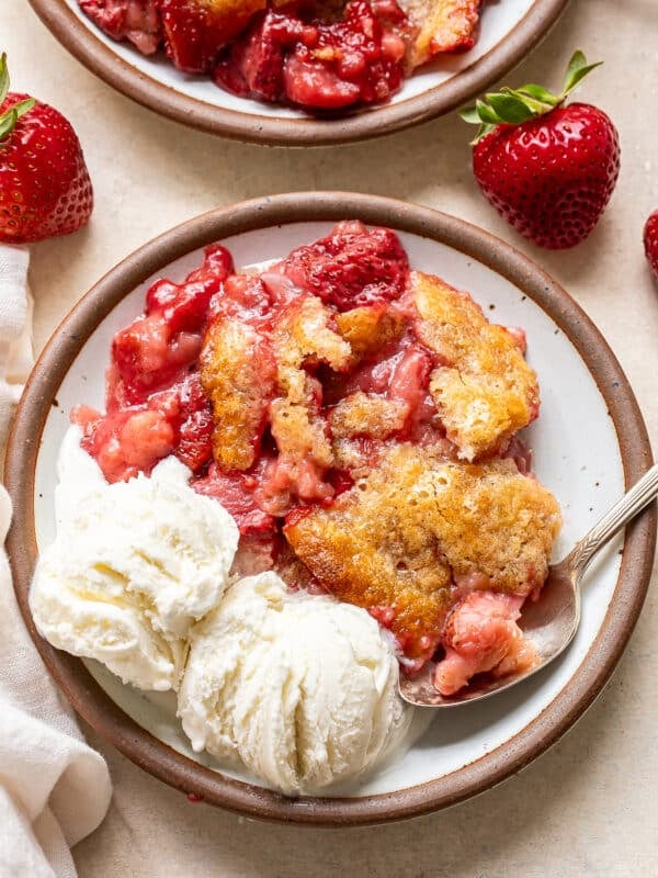 A plate with strawberry cobbler topped with a golden crust, served alongside two scoops of vanilla ice cream. Fresh strawberries are on the table as garnish. A spoon rests on the plate.