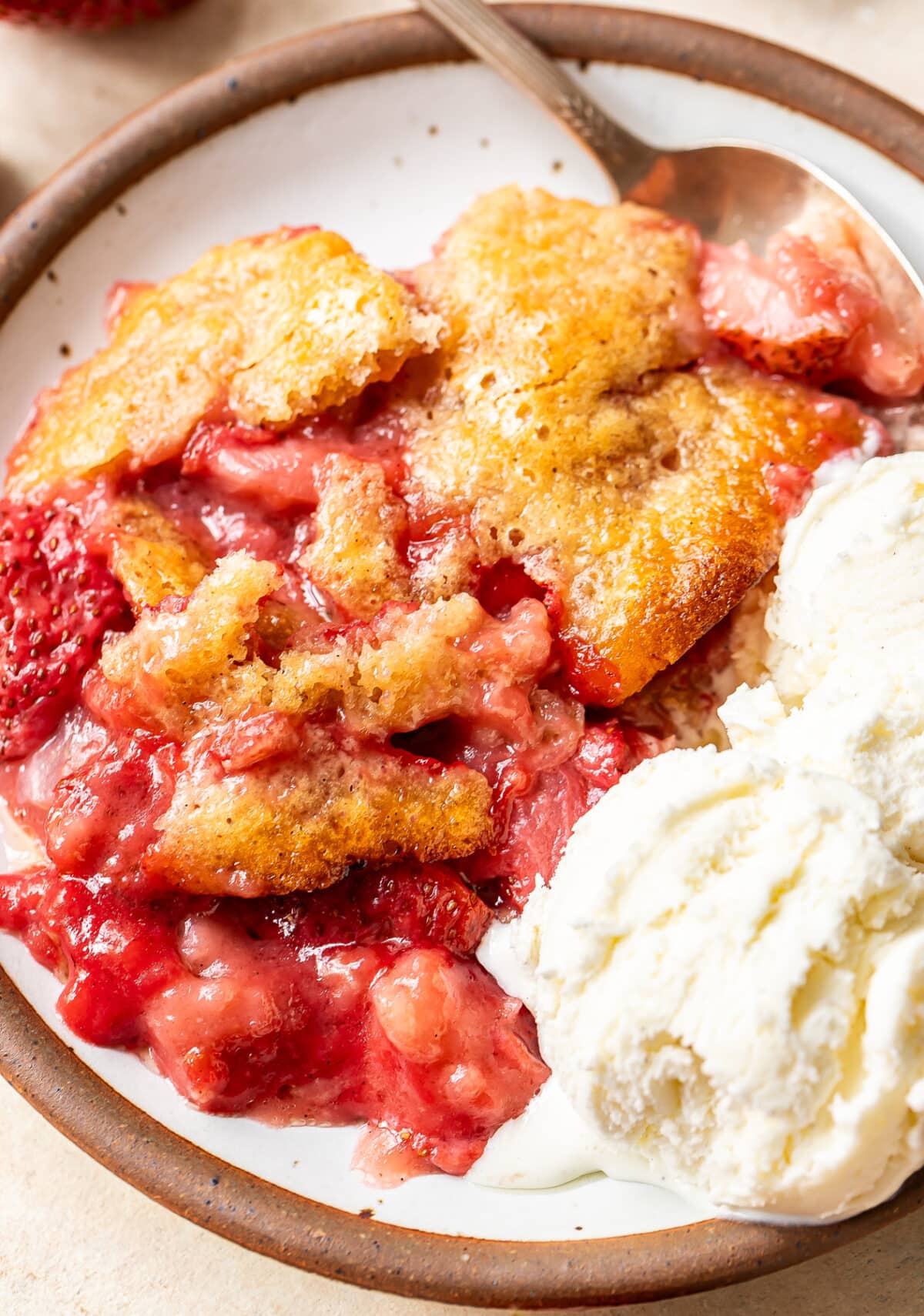 A close-up of a plate with strawberry cobbler, featuring golden baked topping and juicy strawberries, served alongside a scoop of vanilla ice cream with a spoon.
