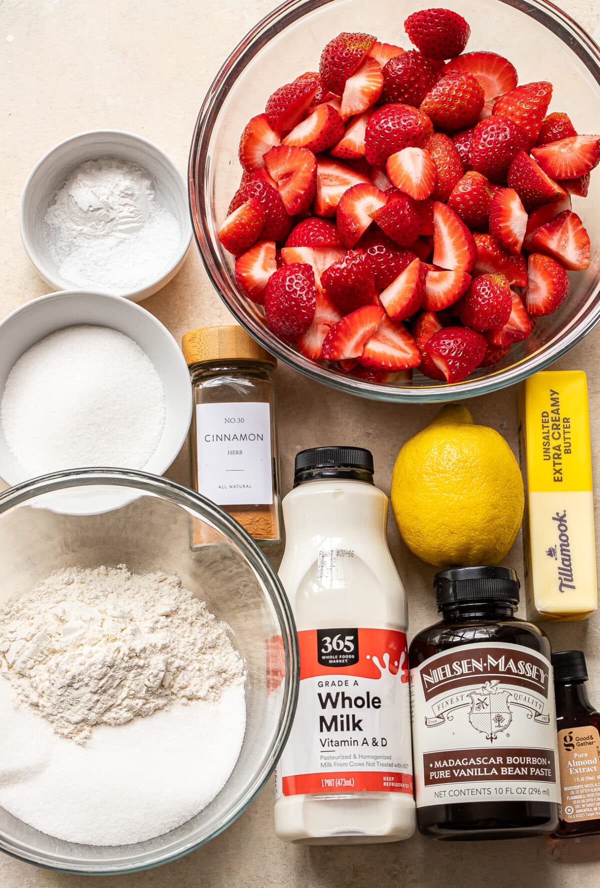 A top-down view of baking ingredients: sliced strawberries, sugar, flour, cinnamon, whole milk, a lemon, vanilla extract, a stick of butter, and a bowl of powdered sugar arranged on a countertop.