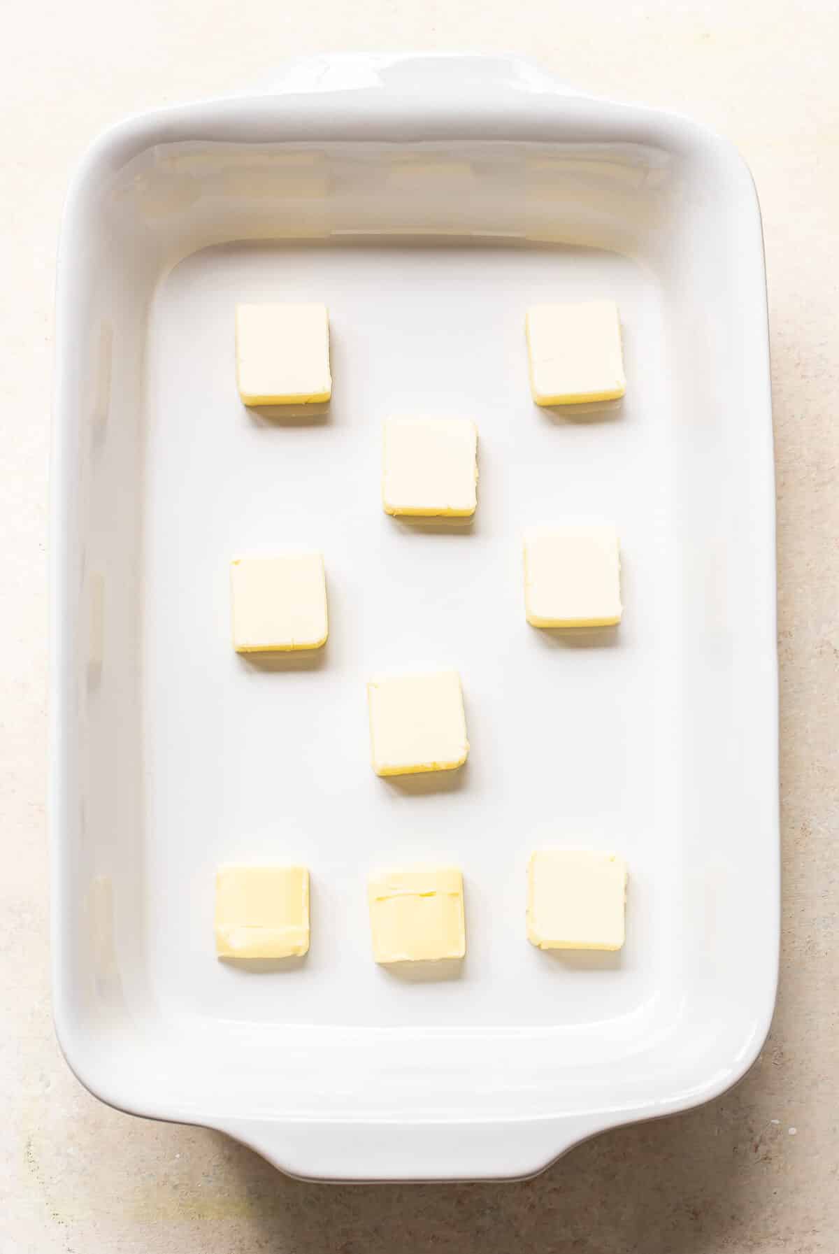 Nine evenly spaced cubes of butter arranged in a grid pattern inside a white rectangular ceramic baking dish on a light-colored surface.