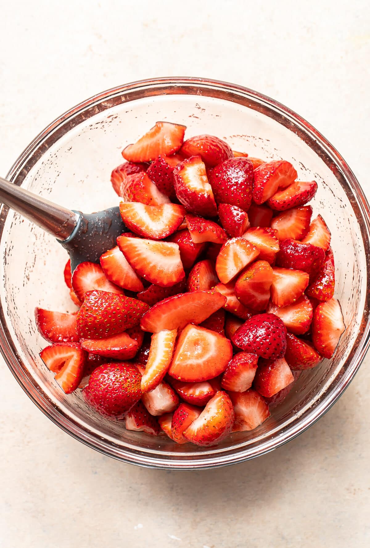 A glass bowl filled with sliced strawberries, with a spatula resting inside, on a light-colored surface.