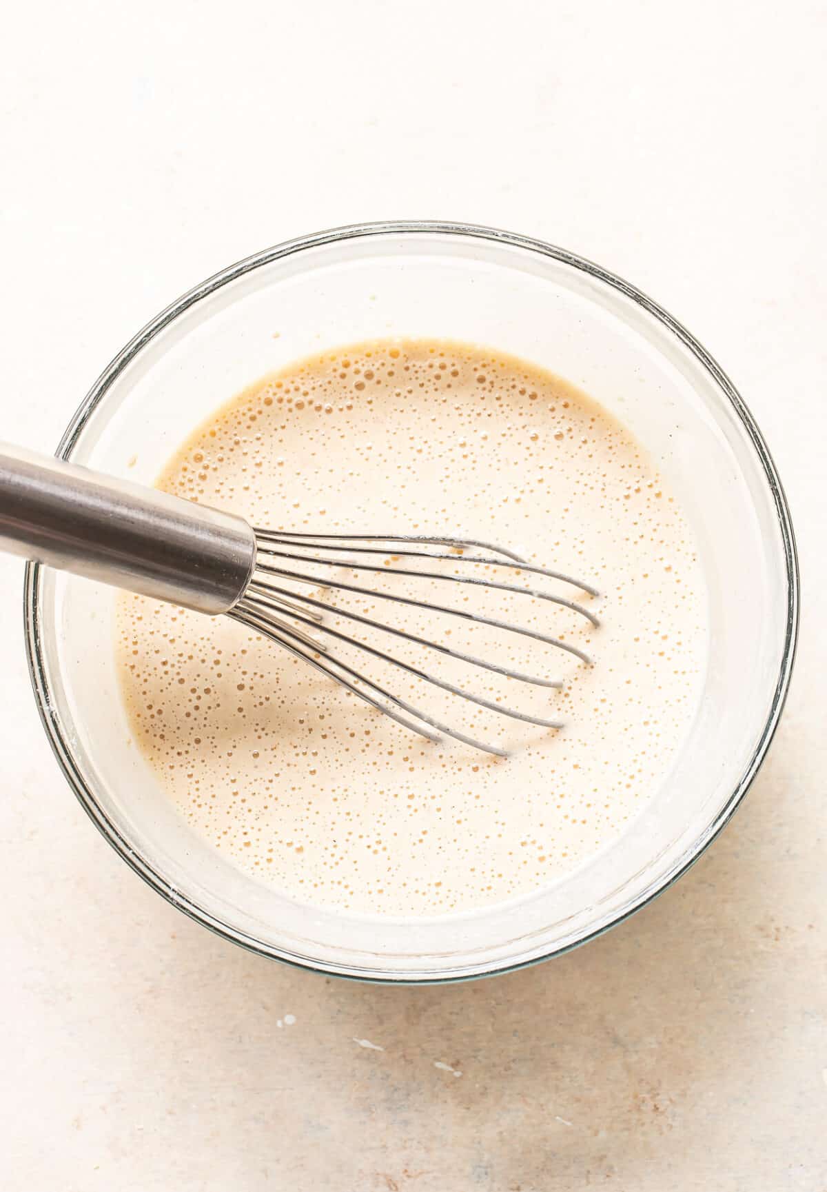 A metal whisk rests in a glass bowl filled with a light, frothy batter or mixture on a pale surface.