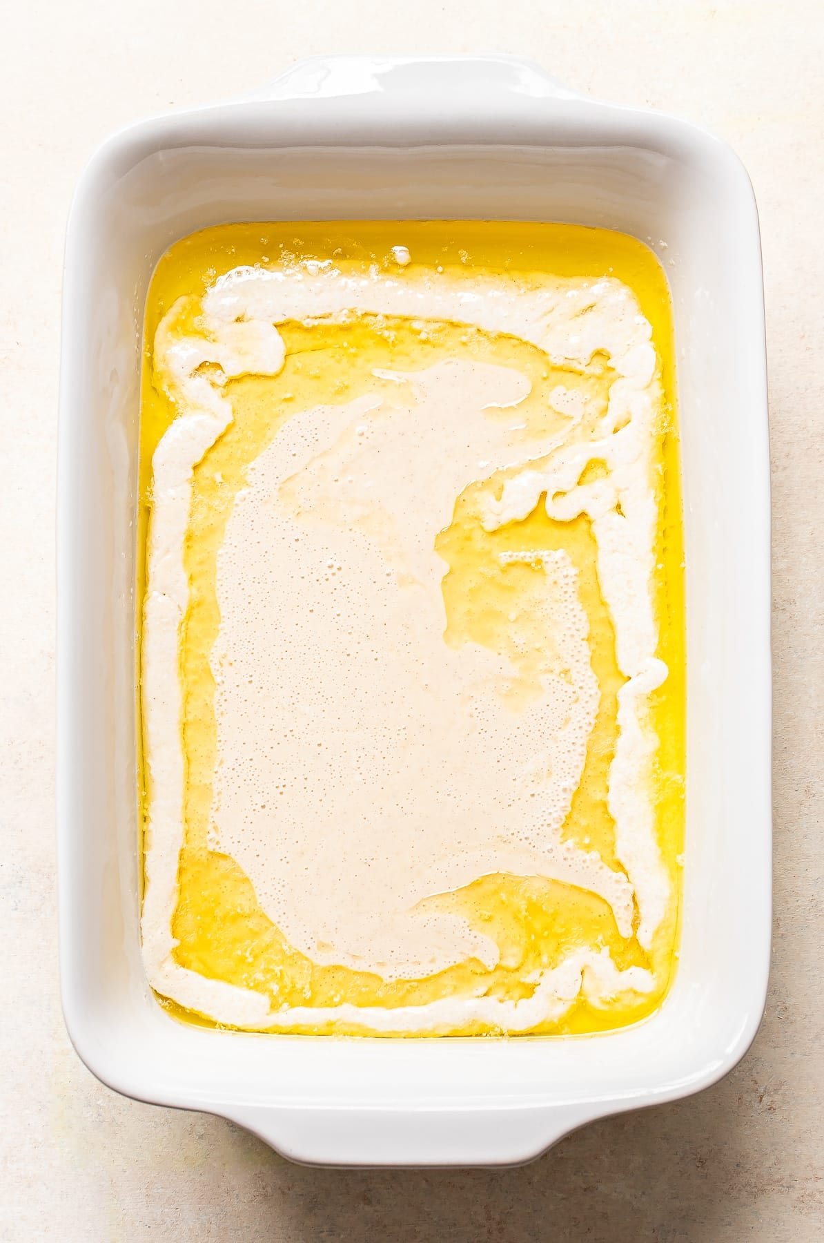 A white baking dish containing melted butter with a layer of batter poured unevenly on top, creating swirled patterns.