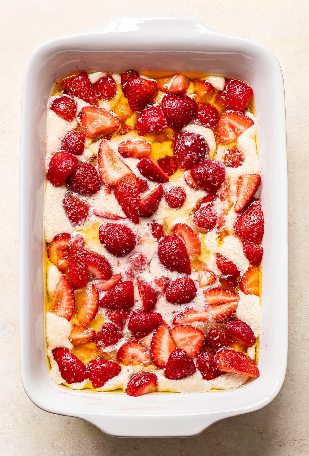 A white baking dish filled with a mixture of sliced strawberries, halved strawberries, and dough, ready to be baked, with a creamy liquid poured over the top.