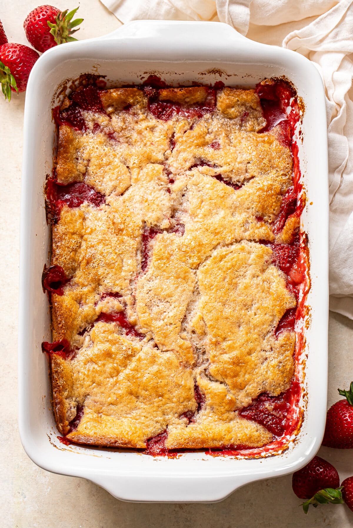 A rectangular white baking dish filled with a golden-brown strawberry cobbler, with juicy strawberries visible around the edges. Fresh strawberries and a white cloth are placed beside the dish.