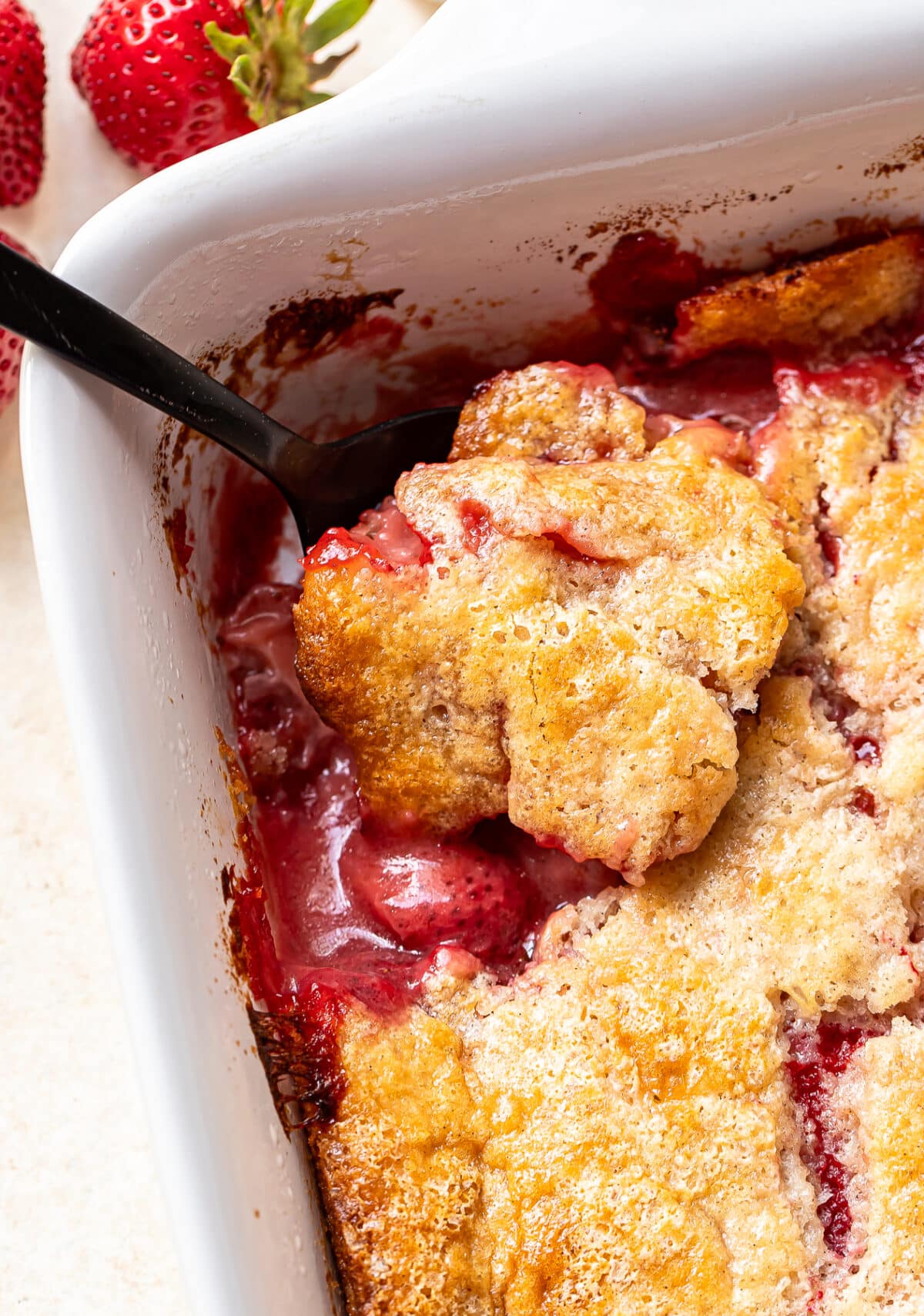 A close-up of a baked strawberry cobbler in a white dish, with a spoon scooping out a portion. The golden, crumbly topping contrasts with the bright red strawberry filling. Fresh strawberries are visible in the background.