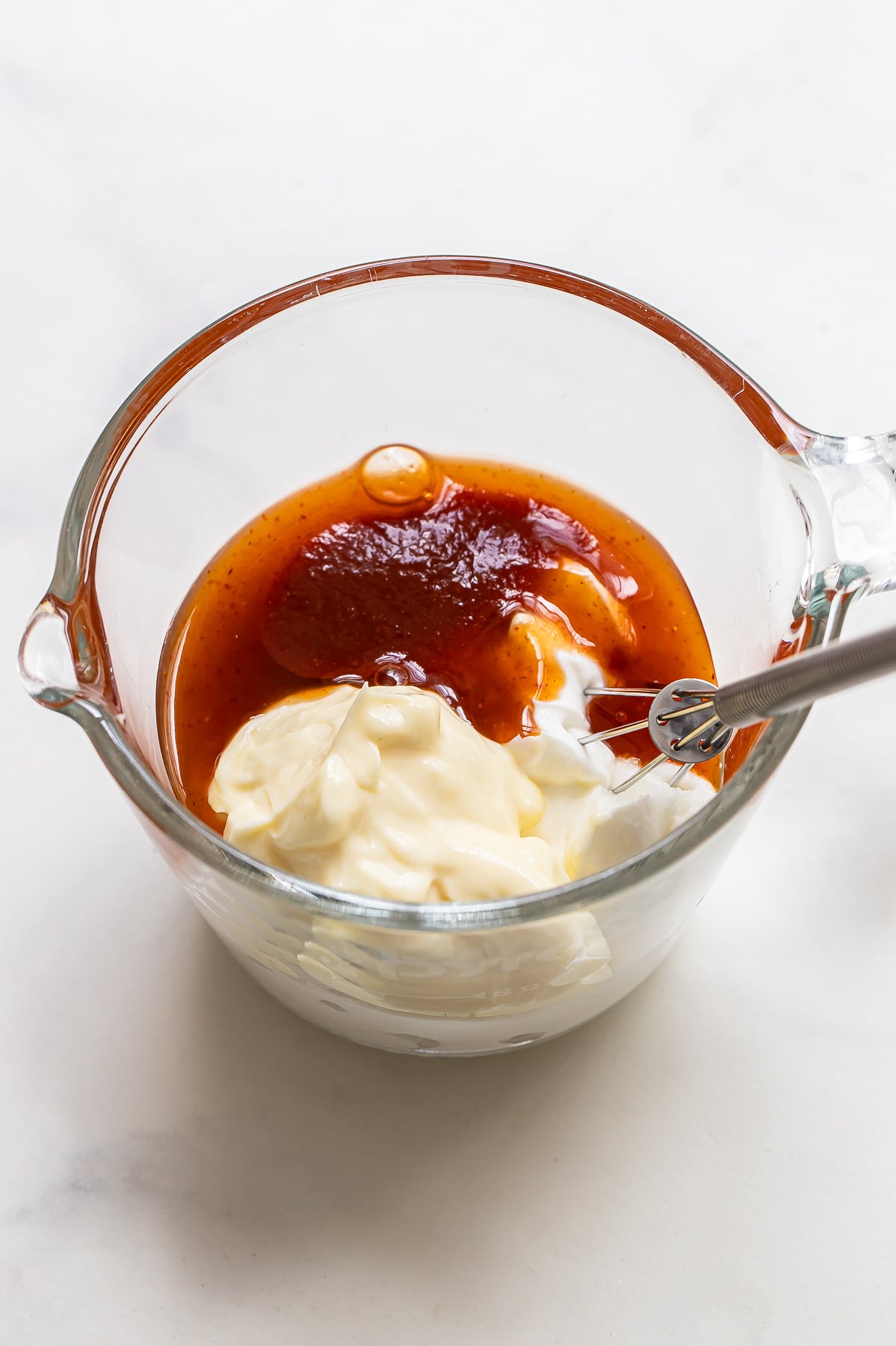 A glass measuring cup containing mayonnaise, a reddish-brown Bang Bang Sauce, and a spoon, ready to be mixed.
