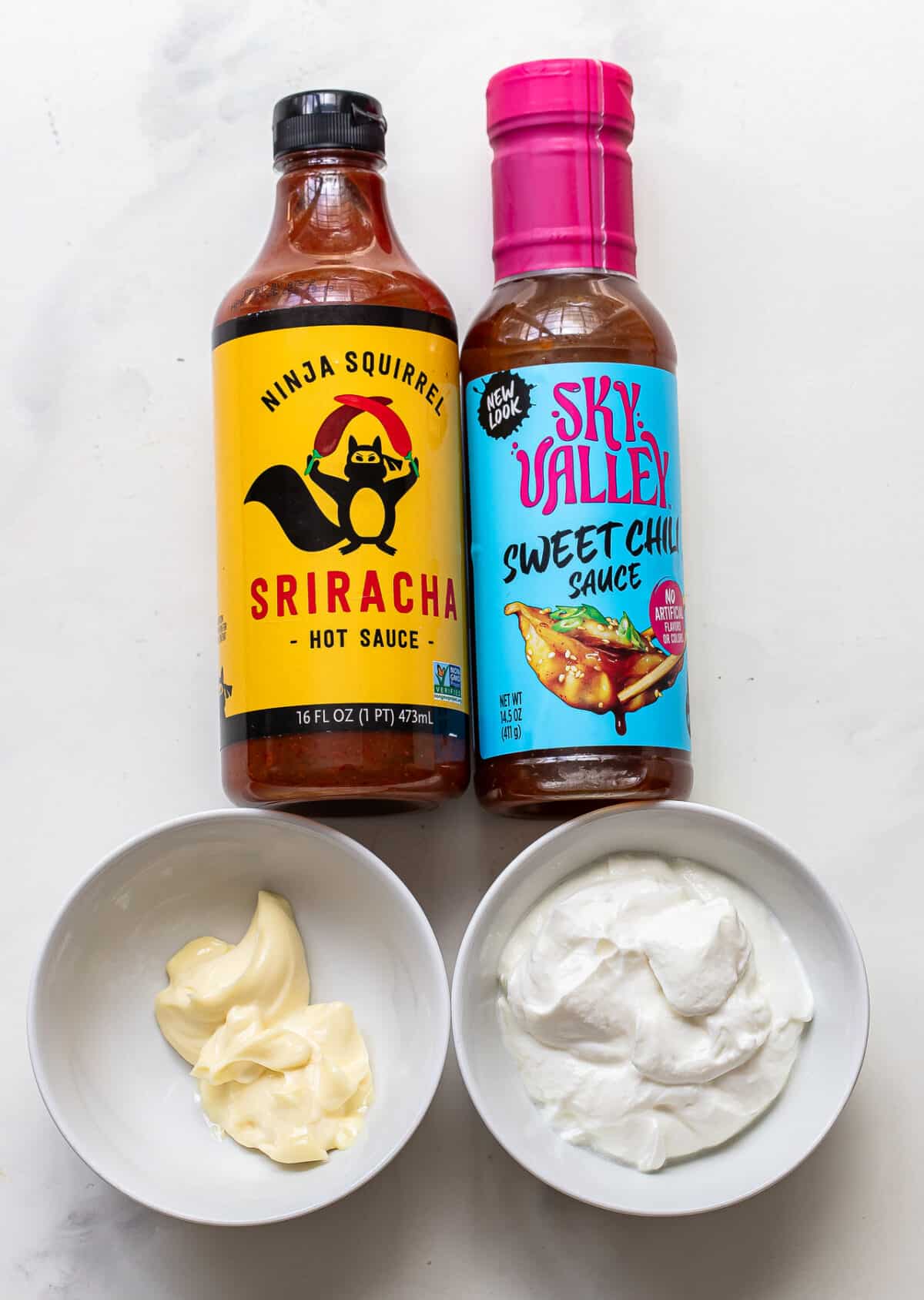 Two bottles of sauce—Sriracha and Sweet Chili—are shown beside two small bowls, one containing mayonnaise and the other containing sour cream or yogurt, ready to be mixed together for a delicious Bang Bang Sauce, all displayed on a white surface.