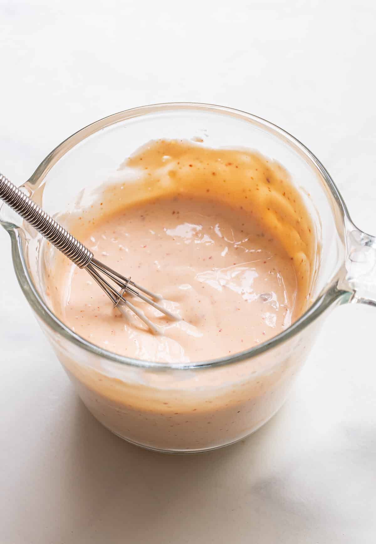 A glass measuring cup filled with creamy, light orange Bang Bang Sauce is being whisked with a metal whisk. The cup sits on a white surface.