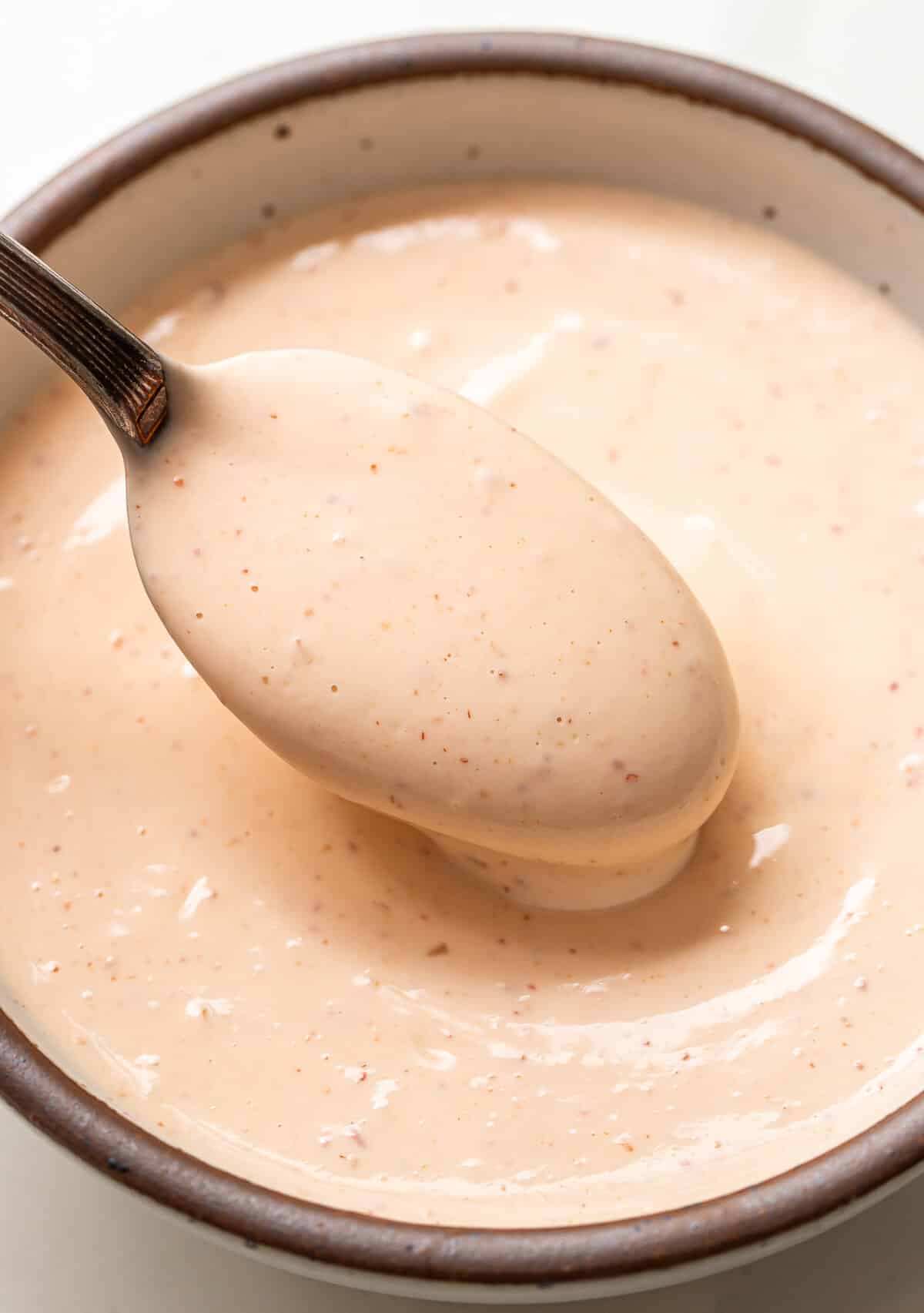 A spoon scoops up creamy, light orange Bang Bang Sauce with specks, from a ceramic bowl.
